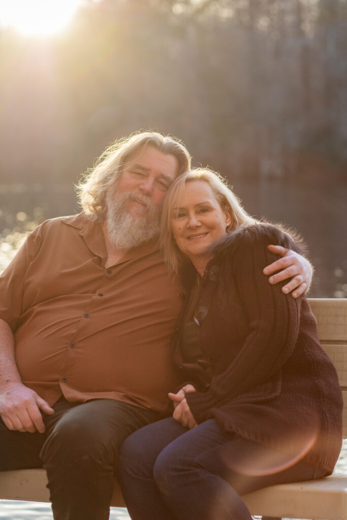 Older couple sitting on a dock hugging lovingly, on a Private Property in Suffolk Virginia, at sunset in November with the golden hour light reflecting off the river behind them.